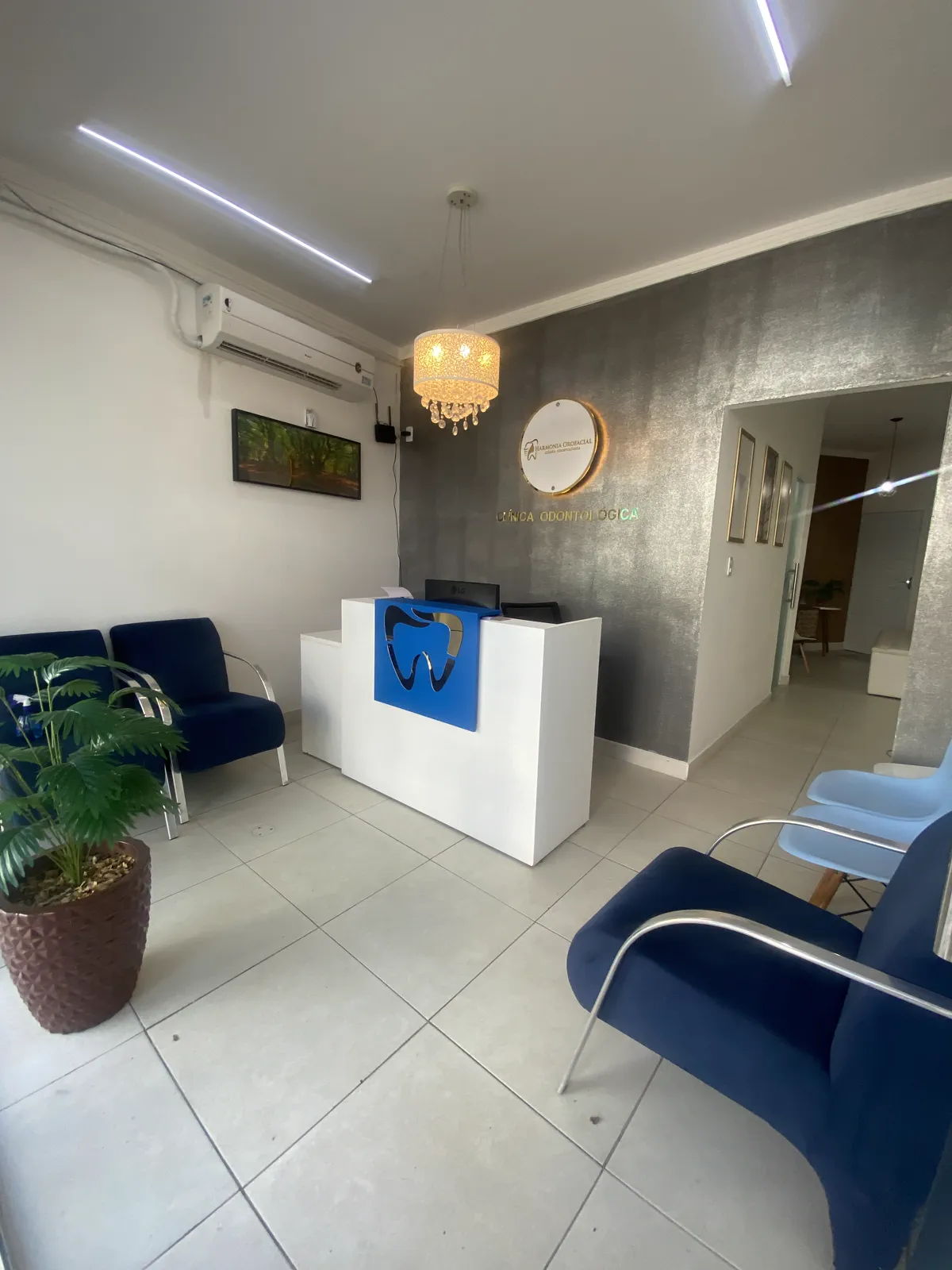 Vista ampla da recepção, com o balcão branco, o lustre aceso e as poltronas azuis